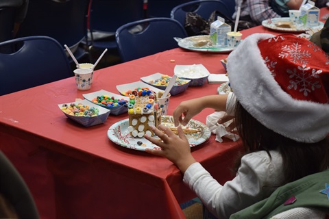 young girl wearing a Santa hat sitting at a table decorating a gingerbread house with cereal and marshmallows